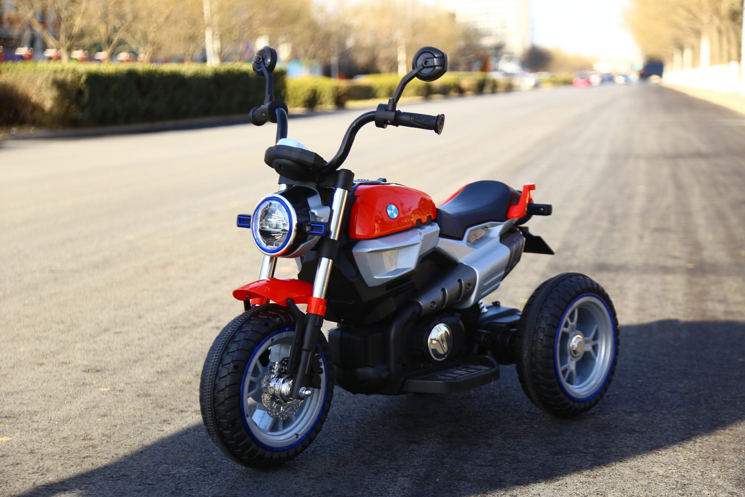 Red and black 12v battery-powered kid’s motorcycle with three wheels parked on an asphalt road outdoors.