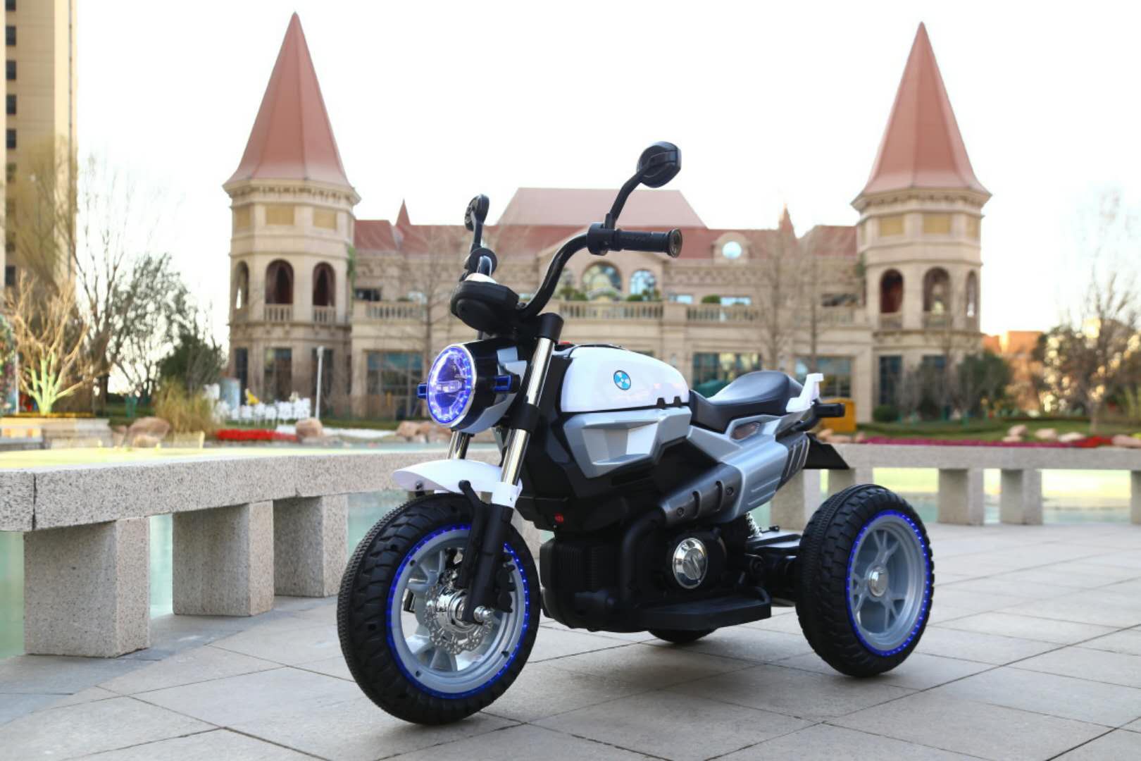 Grey 12V electric ride-on motorbike for kids with LED lights, three wheels, and battery-powered design outdoors near castle background