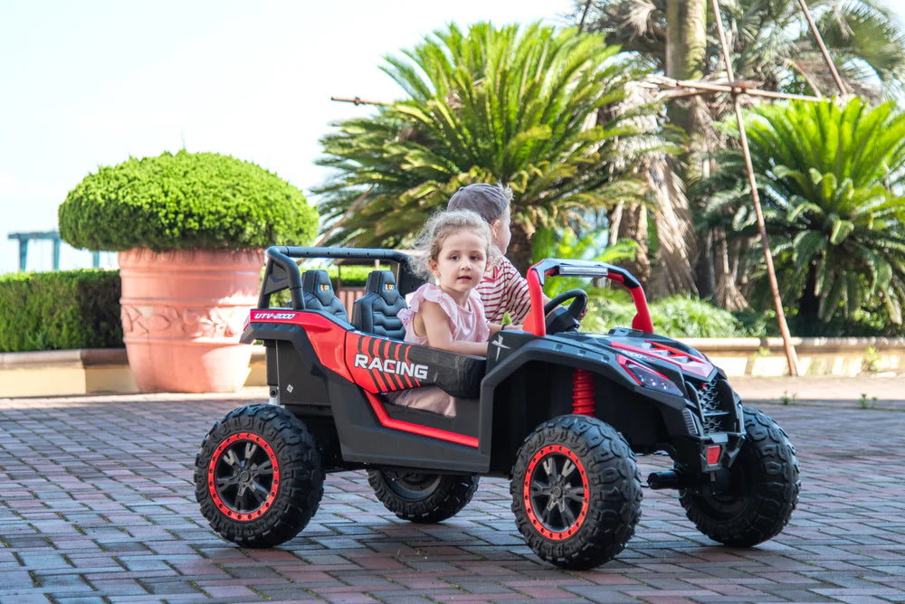 Two children riding a red Stinger Buggy XL 24V 4x4 kids’ ride-on car outdoors on a paved path.