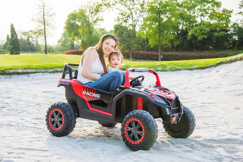 Mother and child sitting in a red and black Stinger Buggy XL 24V 4×4 ride-on car on sandy terrain outdoors