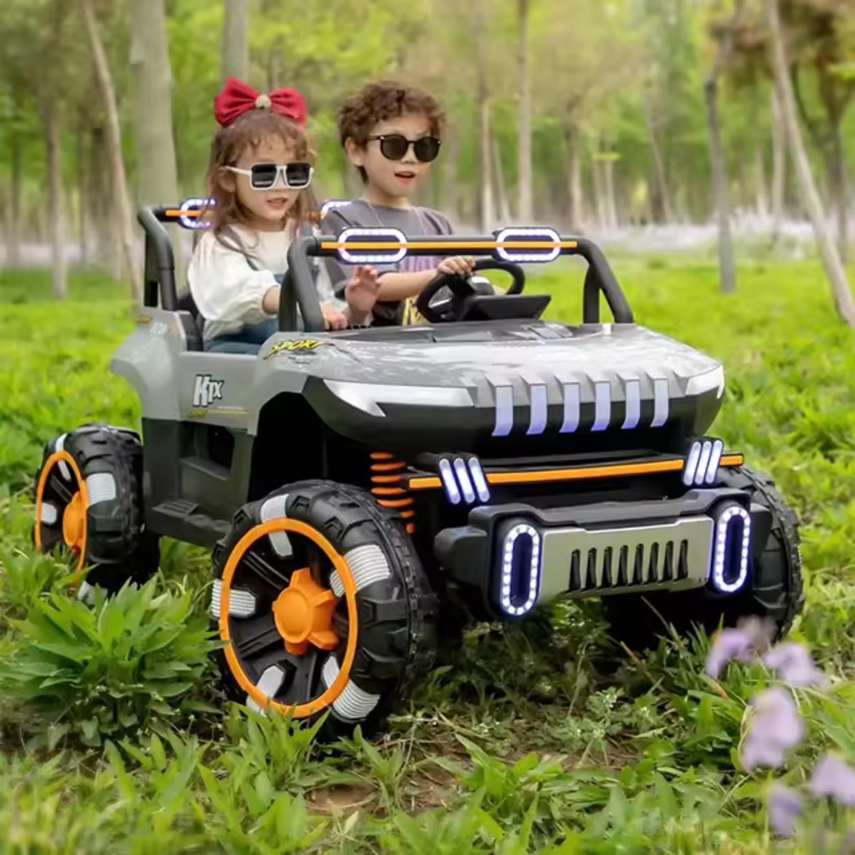 Two kids wearing sunglasses riding a grey 24V 2-seater UTV ride-on buggy on grass outdoors
