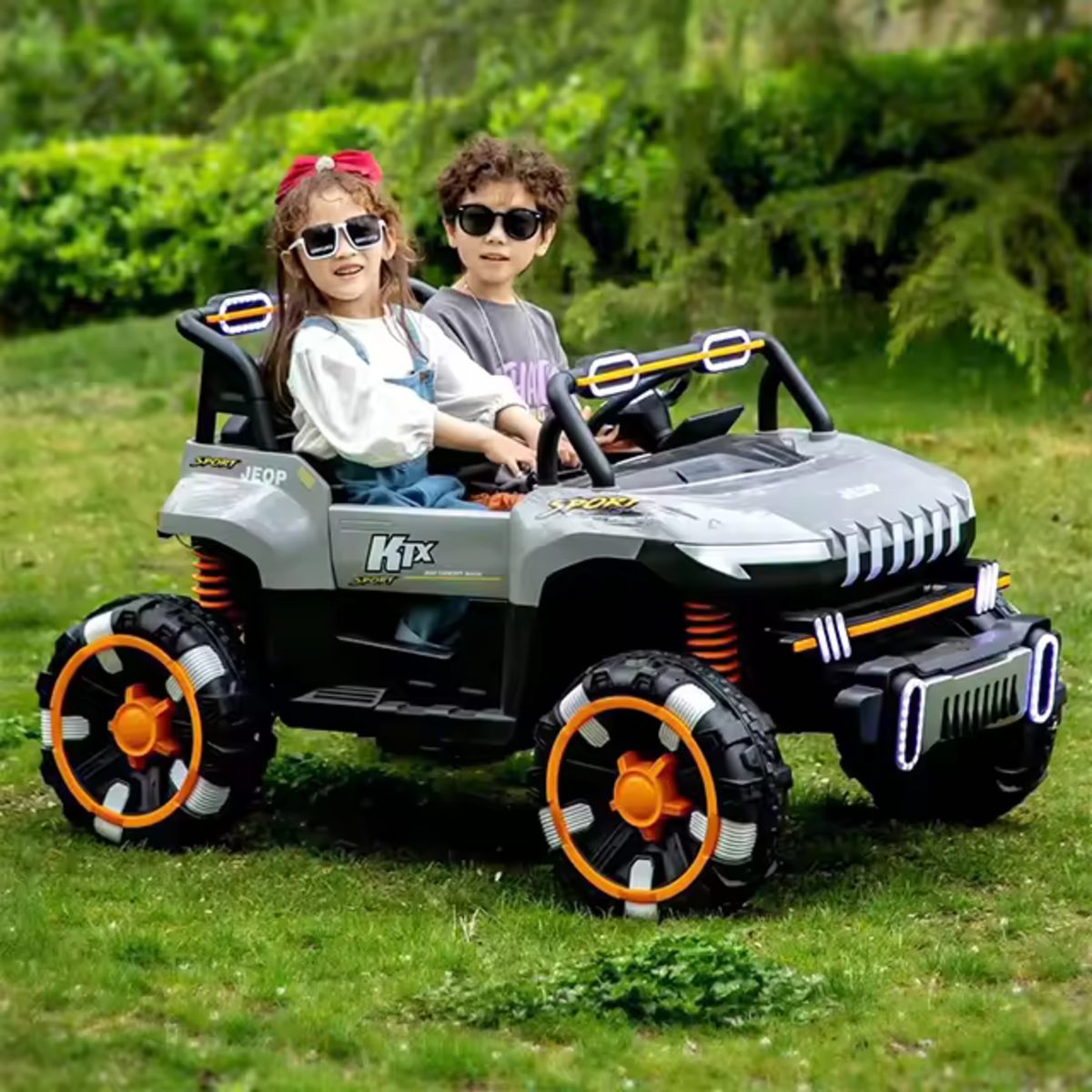 Two kids riding a grey 24V 2-seater UTV ride-on buggy with orange accents on grass outdoors